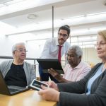 An instructor is standing as he meets with three older adult learners seated in a classroom while they look at their tablets during the class.